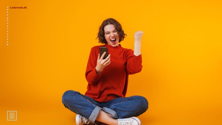 Kvinde, der sidder i skrædderstilling på gulvet, kigger på sin mobiltelefon og laver en vinderbevægelse med hånden.