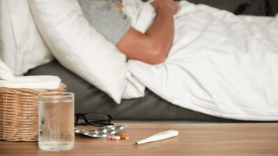 en mand ligger i en sofa med dyne på. i forgrunden ligger et termometer og nogle piller